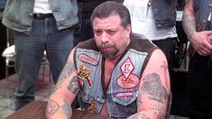 a man sitting at a table with tattoos on his arms and chest, in front of other men