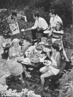 black and white photograph of people sitting around a picnic table in the garden eating food