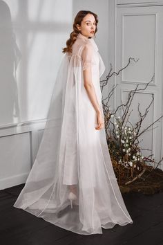 a woman standing in front of a white wall wearing a long veil and dress with sheer sleeves