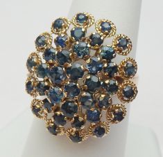 "This is a lovely mid-century sapphire cluster ring, probably made in South East Asia, in the style of a Thai Princess ring. The ring features a cluster of 24 round cut blue sapphires in the center surrounded by a halo of 14 round cut blue sapphires that has rings of twisted gold around them. The ring is just shy of 1\" long on the finger and sits up 0.50\" off the finger. The band is a plain flat style that is 3 mm wide. The ring has a stamp, but it is unreadable. We have tested the ring and it Thai Princess Ring, Vintage Multi-stone Oval Sapphire Ring, Vintage Oval Multi-stone Sapphire Ring, Vintage Blue Cluster Ring With Prong Setting, Vintage Blue Oval Sapphire Ring, Vintage Gemstone Cluster Ring, Vintage Blue Cluster Rings, Vintage Blue Cluster Sapphire Ring, Vintage Blue Sapphire Cluster Ring