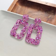 Our Margot Earrings are a bold statement with a hint of glamorous flair. The acrylic rectangle earrings add contemporary style to any outfit, while the matching posts and charms will make you feel effortlessly chic. Hypoallergenic stainless steel posts 15mm stud size Lightweight and durable pink glitter acrylic Trendy Pink Glitter Earrings, Trendy Square Earrings As Gift, Purple Rectangular Jewelry For Party, Pink Glitter Earrings, Modern Pink Rectangular Earrings, Patriotic Bows, Rose Leggings, Rectangle Earrings, Glitter Acrylic