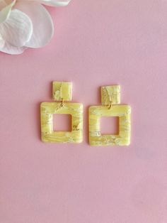 two square shaped yellow earrings sitting on top of a pink surface next to a flower