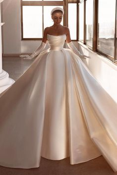 a woman in a white wedding dress standing by a window with her hands on her hips