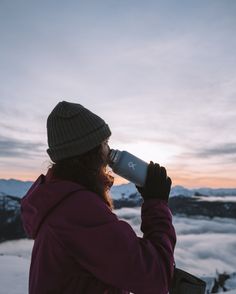 a woman drinking out of a cup while standing on top of a snow covered mountain