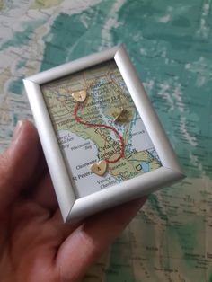 a hand holding a small white frame with a map in the middle and two gold hearts on it