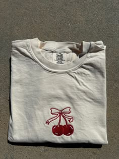 a white t - shirt with cherries embroidered on the front and back, sitting on concrete