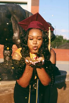 a woman wearing a graduation cap and gown blowing confetti in front of a statue