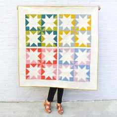 a woman holding up a colorful quilt on a brick wall in front of a white building