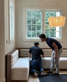 two men working on a couch in a living room next to a window and rug