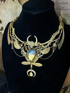 Embrace the mystique of the night sky with this enchanting Celestial Moon Goddess necklace. A true statement piece, this handcrafted treasure marries Art Nouveau elegance with ethereal fantasy elements. Key features: * Intricate gold-toned metalwork featuring delicate leaves, fans, and celestial motifs * Stunning centerpiece labradorite stone, showcasing mesmerizing flashes of blue and gold * Crescent moon shapes echoed throughout the design * Multiple layers of chains and decorative elements fo Fantasy Brass Jewelry For Gifts, Moon Phase Spiritual Jewelry, Fantasy Moon Shaped Jewelry Gift, Handmade Celestial Labradorite Necklaces, Gold Fantasy Necklace In Metal, Handmade Celestial Labradorite Necklace, Handmade Labradorite Celestial Necklace, Unique Handmade Moon Shaped Necklace, Handmade Moon Shaped Necklace