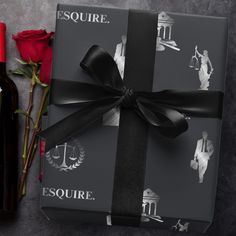 a gift wrapped in black paper next to a bottle of wine and rose buds on a table