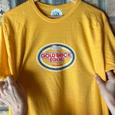 a person holding up a yellow t - shirt with an old brick eggs logo on it