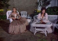 two women sitting on white chairs in front of a brick wall with yellow flowers and potted plants