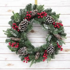 a christmas wreath with pine cones and red berries hanging on a white wooden wall next to a candle