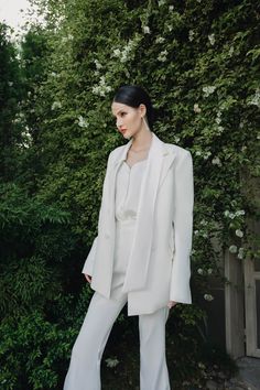 a woman standing in front of a bush wearing white pants and a jacket with long sleeves