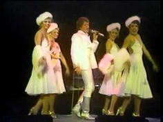 the four women are dressed in white outfits and singing on stage with one woman holding a microphone