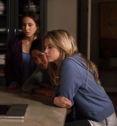 three women looking at a laptop computer screen in a kitchen with one woman leaning on the counter