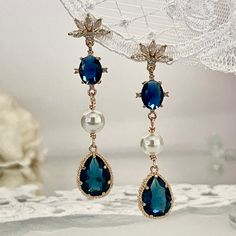 Dive into the elegance of the past with our Navy Blue Art Deco Earrings, a perfect blend of Art Nouveau charm and Bohemian chic. Each earring begins with a beautifully detailed floral post, encrusted with clear zirconia crystals, leading to a mesmerizing navy oval crystal, surrounded by four petite zirconias. Below, a delicate white crystal pearl adds a touch of romantic Coquette flair, trendy for both everyday fashion and chic boho weddings. The pièce de résistance is the navy blue crystal tear Blue Clip-on Earrings For Wedding, Blue Dangle Chandelier Earrings For Anniversary, Blue Chandelier Drop Earrings For Anniversary, Formal Blue Pearl Drop Earrings, Blue Teardrop Pearl Earrings, Elegant Blue Earrings For Party, Formal Blue Dangle Bridal Earrings, Blue Pearl Drop Earrings For Formal Occasions, Blue Teardrop Chandelier Earrings For Wedding