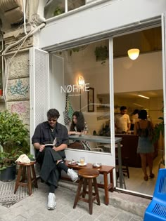 a man sitting on a bench in front of a store window reading a book and drinking coffee