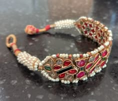 "Here is an awesome antique Indian bracelet or bazuband.  It is 22k yellow gold and the wire that runs on the edges is silver.  The backside is enameled in a green, red and white flower pattern.  The center of the bracelet is comprised of nine separate panels that are kundan set with natural and untreated rubies, emeralds and diamonds.  The gemstones are all old table or flat cuts.  There are seed pearls strung along the edges that culminate in tightly spiraled coil of pearls and then into fabri Bajubandh Design Gold, Traditional Ruby Bangle With Stone Work, Ruby Stone Bangles Gold, Ruby Emerald Bangles, Ruby Bangle With Intricate Design, Luxury Traditional Ruby Bracelets, Gold And Pearl Bracelet, Indian Bracelets, Antique Gold Bracelet