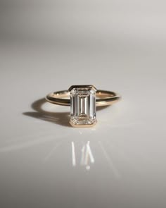 an emerald - cut diamond ring sits on a white surface in front of the camera