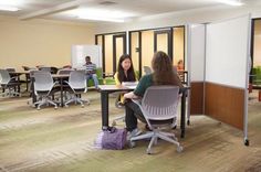 two women sitting at desks in an office setting with whiteboards on the walls