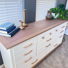 a white dresser with drawers and books on top, next to a potted plant