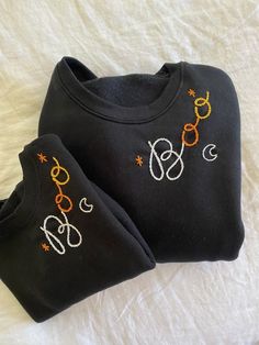 two black sweatshirts with embroidered letters on them sitting on a white bed spreader