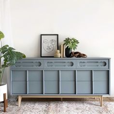 a blue dresser with two plants on top and a framed portrait above it in a white room