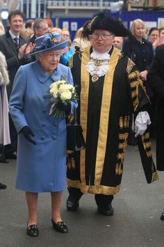 an older woman in a blue coat and hat walking next to an older man wearing a black suit