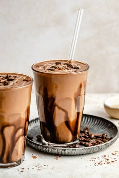 two glasses filled with chocolate milkshakes sitting on top of a plate next to coffee beans