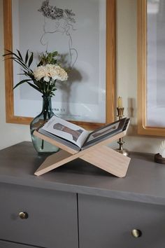 a vase with flowers and an open book on top of a dresser next to two framed pictures