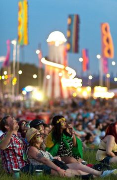 many people are sitting in the grass at an outdoor concert with bright lights behind them