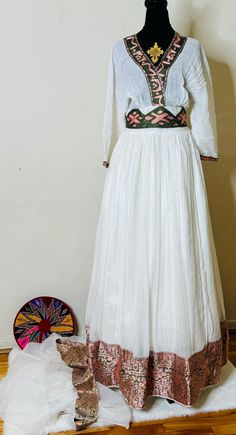 Ethiopian and Eritrean traditional dress (habesha kemis) outfit. The attire consists of a white long-sleeved top with intricate embroidery around the neckline and waist, as well as a full-length white skirt with matching embroidery near the hem. The embroidery features pink and green patterns, adding elegance and contrast to the white fabric. beautiful dress.elegant dress Eritrean Clothing, Matching Embroidery, Green Patterns, Ethiopian Dress, Habesha Kemis, Intricate Embroidery, Oct 11, White Skirt, Silver Spring