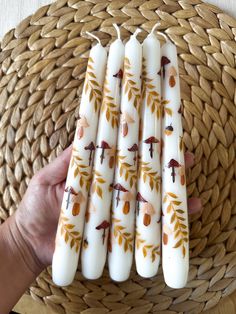 four white candles with leaves and birds on them are held in front of a woven wicker basket