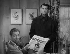 two men sitting at a table with a newspaper in front of them, one reading the paper