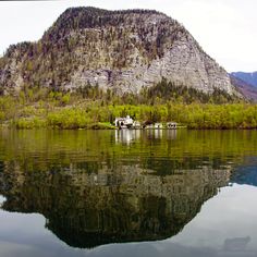 a house on the shore of a lake in front of a large mountain with green trees