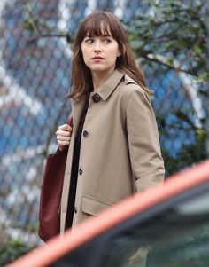 a woman in a trench coat is looking at the camera while holding her purse and walking