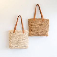 two straw bags hanging up against a white wall with leather handles and straps on each side