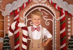 a young boy wearing suspenders and a bow tie holding a candy cane in front of a gingerbread house