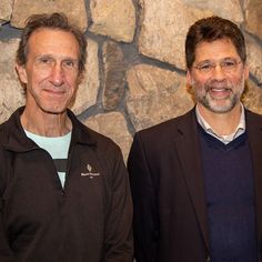 two men standing next to each other in front of a stone wall with rocks on it