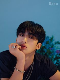 a young man with black hair wearing a necklace and bracelet sitting in front of a blue wall