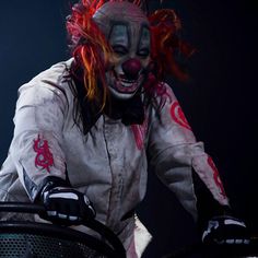 a clown with red hair and makeup is riding on a metal object in the dark