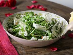 a white bowl filled with green salad on top of a wooden table next to red petals