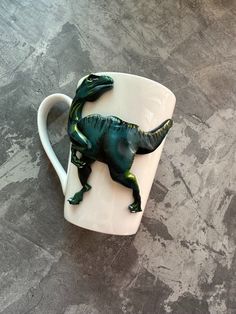 a green toy dinosaur sitting on top of a white coffee cup in the shape of a t - rex