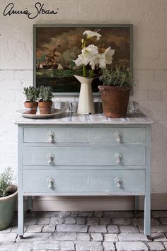 a blue dresser with potted plants on top and an old painting above it in the background