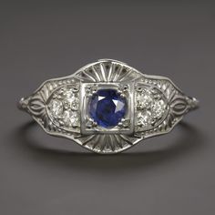 This beautiful and unique ring features a richl blue sapphire set in a diamond studded vintage style design that will never go out of style! The sapphire has a richly saturated blue hue that contrasts strikingly with the 14k white gold setting. Utterly classic and romantic, the antique style setting is richly textured with intricate filigree and milgrain. The effect is chic but also dreamy and softly feminine, a perfect choice for a timelessly stylish right hand ring or alternative engagement ri Classic Sapphire Halo Ring, Classic Sapphire Birthstone Ring, Art Deco Blue Diamond Ring With Accents, Art Deco Blue Diamond Ring With Center Stone, Art Deco Blue Sapphire Ring With Center Stone, Blue Art Deco Diamond Ring With Center Stone, Classic Royal Blue Round Sapphire Ring, Blue Art Deco Diamond Ring With Accents, Classic Blue Sapphire Ring In 14k White Gold