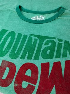 What Are You Waiting For? Just Dew It! Mountain Dew's logo may have changed several times since the 1940's, but that crisp and refreshing taste has always remained. Whether you're a hardcore gamer or just enjoy a cold Dew, show your love with this authentic licensed Mountain Dew t-shirt! We picked one of Mt. Dew's longest lasting logos and gave it a fun, lightly distressed look. To complete the retro feel, we’ve printed it on the highest quality poly-cotton Kelly Green ringer t-shirt around. Thi Green Tops With Graphic Design For Fan Merchandise, Mountain Dew Logo, Mt Dew, Ringer Shirt, Retro Mountain, Mens Fade, Mountain Dew, Ringer Tee, Mens Big And Tall