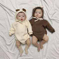 two baby babies laying on top of a white bed next to each other wearing bear onesuits