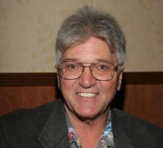 a man wearing glasses and a gray jacket smiles at the camera in front of a wall